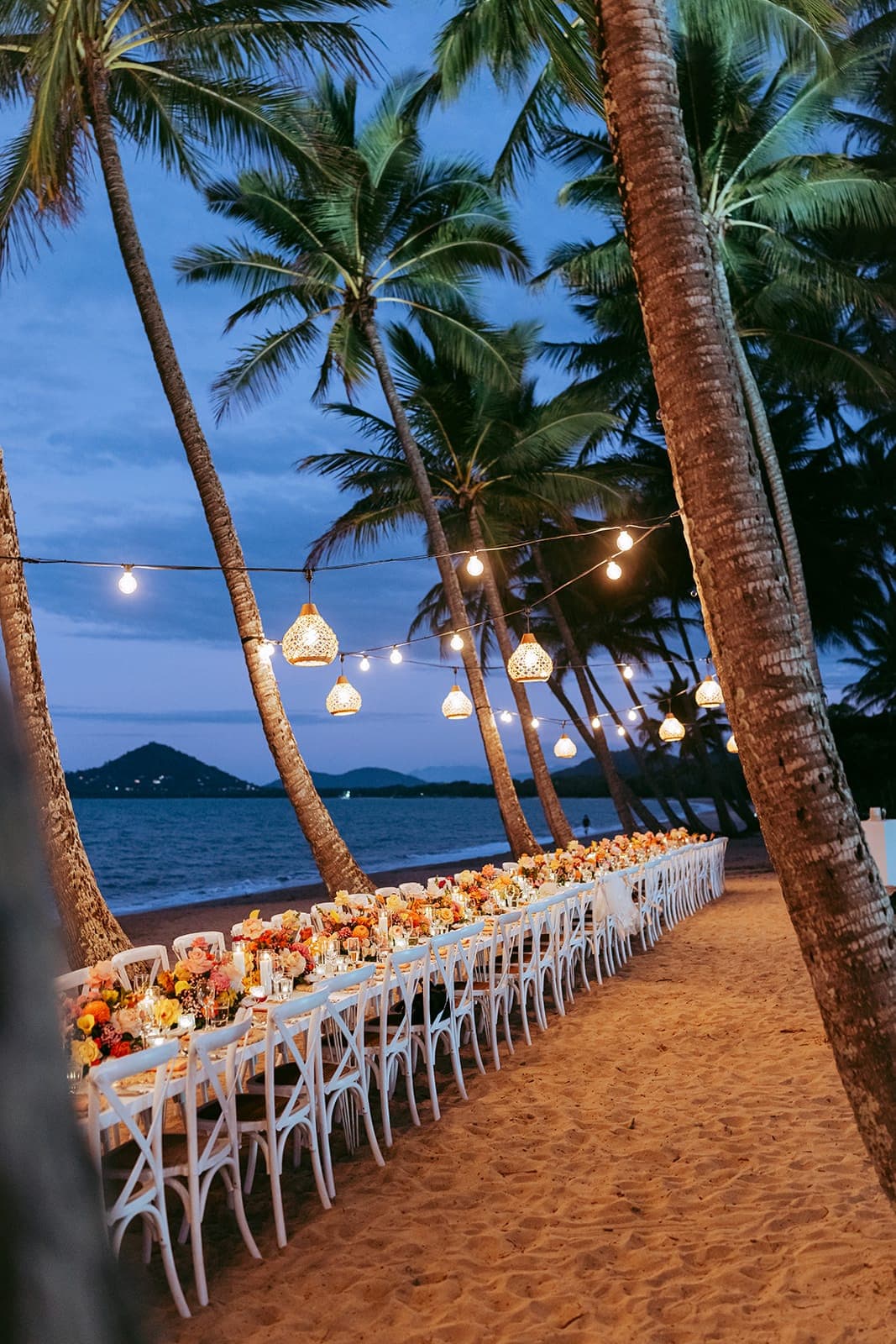 A long table set on the sand at blue hour under coconut palms strung with lanterns, pink, orange and yellow flowers down its length, the Coral Sea behind